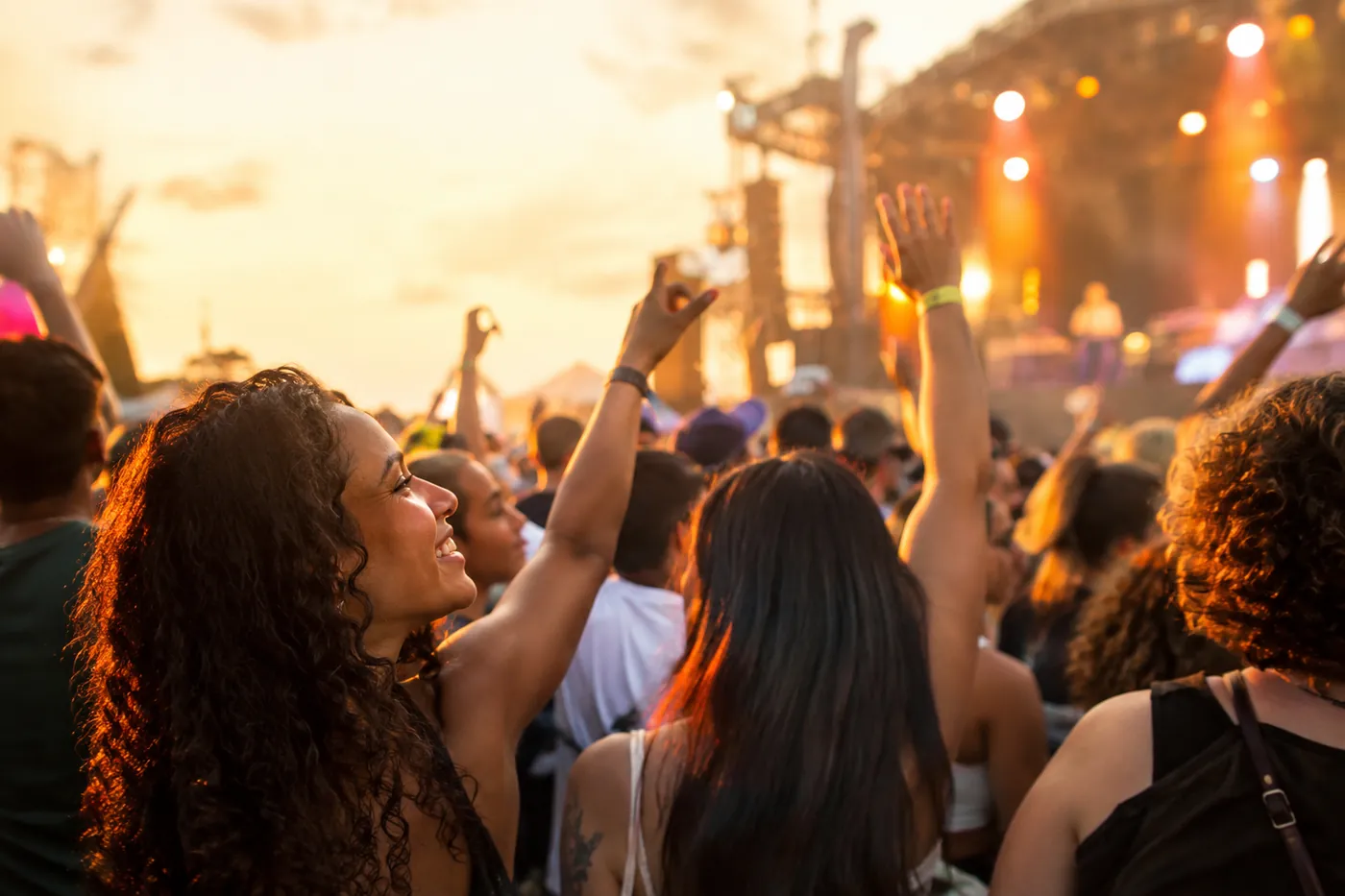 Público em um evento ao ar livre no fim da tarde, de braços levantados diante de um palco iluminado